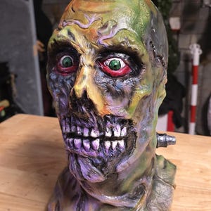 May include: A detailed zombie mask in green, purple, and yellow. The mask has exposed teeth, red-rimmed green eyes, and a metal bolt on the side. A Halloween prop.