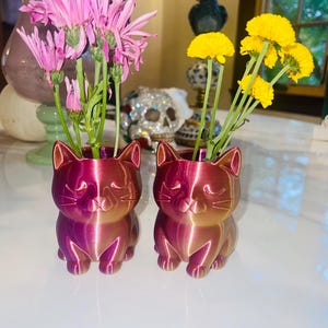 May include: Two cat-shaped vases in a gradient of pink and gold, each holding flowers. One vase has pink flowers, the other yellow. The vases are on a white surface, with decorative items in the background.