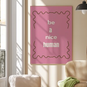 Könnte beinhalten: Ein gerahmtes rosa Poster mit den Worten "be a nice human" in weißer Schrift. Das Poster hat einen braunen, wellenförmigen Rahmen und hängt an einer Wand. Der Raum hat eine neutrale Farbgebung mit einem Stuhl und einem Hocker.