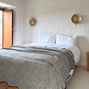Könnte beinhalten: Ein Schlafzimmer mit einem Bett, das mit einer grau-weiß gemusterten Decke bedeckt ist. Zwei weiße Kissen liegen auf dem Bett. Zwei goldfarbene Wandleuchten sind an der weißen Wand über dem Bett angebracht.