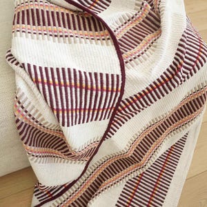 May include: A woven throw blanket featuring a repeating pattern of wide cream-colored stripes alternating with textured bands of deep burgundy, tan, and thin accents of bright orange and yellow, creating a structured and rhythmic visual design.