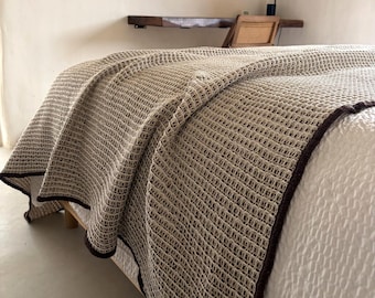 May include: A beige waffle-knit blanket with a dark brown border is draped over a bed. The blanket's texture is visible, and it covers a white bedspread. The bed is made of wood.