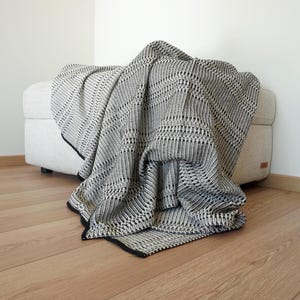 May include: A monochrome woven throw blanket with a geometric pattern, draped over a light-coloured ottoman. The blanket has a black border. The ottoman is neutral-toned and sits on a wooden floor.