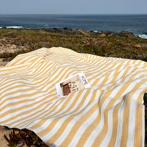 Könnte beinhalten: Eine gelb-weiß gestreifte Decke liegt am Strand in der Nähe des Meeres. Ein Magazin mit dem Wort "HOME" auf dem Cover liegt auf der Decke. Die Decke besteht aus gewebtem Material.
