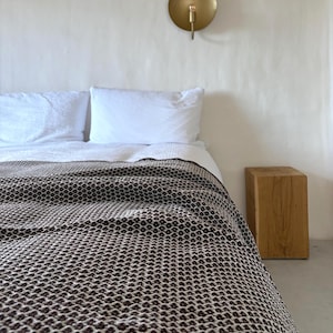 Könnte beinhalten: Ein Bett mit weißer Bettwäsche und einer gemusterten braun-weißen Decke. Zwei weiße Kissen liegen auf dem Bett. Ein kleiner Holzblock befindet sich neben dem Bett. Eine goldfarbene Wandleuchte ist an der weißen Wand angebracht.