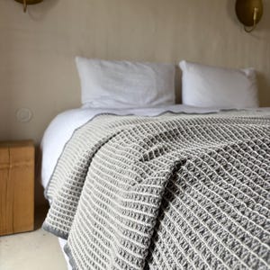 Könnte beinhalten: Eine grau-weiß karierte Strickdecke liegt auf einem Bett mit weißen Kissen. Die Decke hat ein strukturiertes, gitterartiges Muster und einen grauen Rand. Das Bett steht vor einer neutralfarbenen Wand.