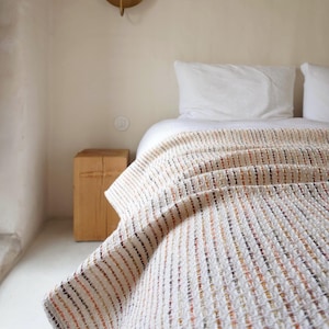 May include: A cream-coloured woven blanket with brown and orange stripes, draped over a bed with white bedding. The room has a minimalist design, with a wooden side table. The blanket's dimensions are not visible.