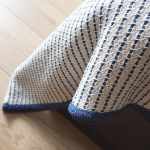 Peut inclure: Un textile tissé présentant un motif texturé de rayures verticales crème et bleu foncé. Le tissu possède une structure de tissage en grille distincte et est fini avec un ourlet bleu foncé uni sur les bords, créant une apparence nette et structurée.
