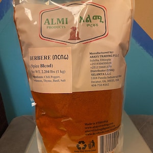 Op de afbeelding: Een transparante plastic zak met ALMI BERBERE kruidenmix. De zak bevat een roodbruin poeder en vermeldt de ingrediënten: chilipeper, kardemom, tijm, basilicum en zout. Het nettogewicht is 1 kg.