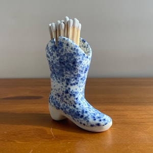 May include: A ceramic match holder shaped like a cowboy boot, decorated with a blue speckled pattern. The boot is filled with wooden matches with white tips. The holder sits on a wooden surface.