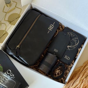 May include: A black gift box containing a black travel bag, passport holder, luggage tag, and a candle. The items have gold accents and the initials "RD-". The passport holder features a world map design.