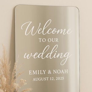 May include: A rectangular mirror sign with rounded corners featuring white text that reads Welcome TO OUR wedding EMILY & NOAH AUGUST 12, 2025. The surface is reflective and smooth, displaying a clear script font for the words Welcome and wedding.