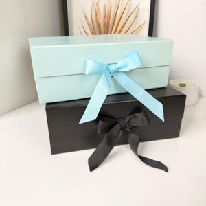 May include: A light blue rectangular gift box with a matching light blue ribbon bow sits stacked on top of a black rectangular gift box featuring a black ribbon bow. Both boxes have a smooth finish and are positioned horizontally on a surface.