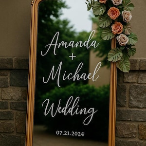 May include: A tall rectangular mirror with a gold frame, featuring white cursive text that reads Amanda + Michael Wedding and the date 07.21.2024. The top right corner is decorated with a cluster of artificial peach roses and green monstera leaves.