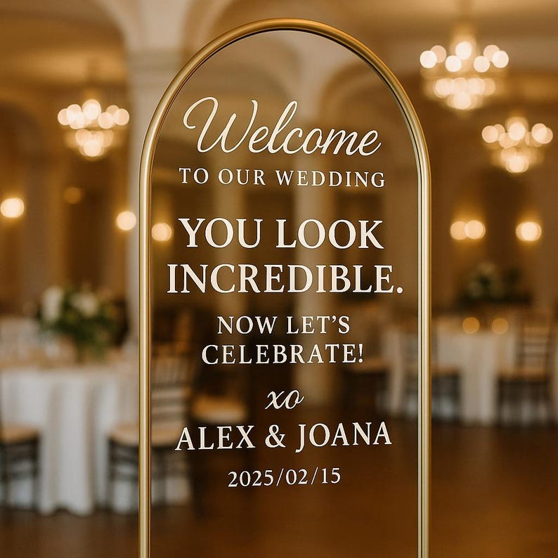 May include: An arched gold metal frame holding a clear acrylic wedding sign with white text that reads: Welcome TO OUR WEDDING YOU LOOK INCREDIBLE. NOW LET'S CELEBRATE! xo ALEX & JOANA 2025/02/15. The sign is freestanding and features a minimalist design.