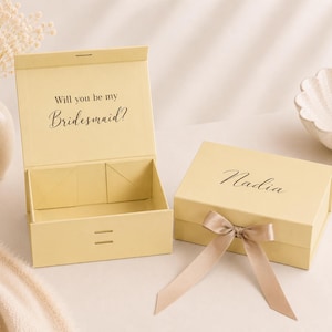 Puede incluir: Dos cajas de regalo rectangulares de color amarillo pálido con cierres magnéticos. Una caja está abierta y muestra el texto impreso en negro "Will you be my Bridesmaid?" en la tapa interior. La otra caja está cerrada, con el nombre "Nadia" y un lazo beige.