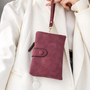 Puede incluir: Una cartera de mano color burdeos profundo con una textura mate similar al ante. La cartera rectangular incluye una solapa con cierre de botón a presión en la parte frontal y una correa de muñeca a juego sujeta a un broche giratorio dorado.