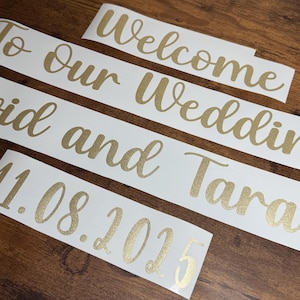 May include: Three white vinyl adhesive decals with gold glitter lettering. The text reads Welcome, To Our Wedding, and David and Tara, followed by the date 11.08.2025. The lettering features a cursive script font style on a smooth white backing.