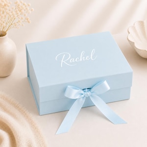 May include: A light blue rectangular gift box with a hinged lid, featuring the name Rachel printed in white script on the top. The box is secured with a matching light blue satin ribbon tied into a bow at the front centre of the lid.