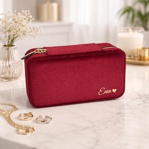 May include: A burgundy velvet jewelry case with a gold zipper and the name "Erin" and a heart symbol embroidered in gold. A gold necklace and two rings are on the surface.