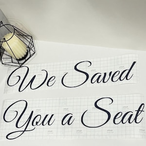 May include: Two sheets of clear transfer tape with a grid pattern, each featuring black vinyl lettering in a cursive script. The top sheet displays the text "We Saved" and the bottom sheet displays the text "You a Seat" with Cricut branding.
