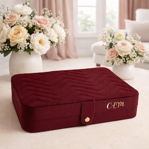 May include: A rectangular, deep red velvet jewellery box featuring a textured chevron pattern on the lid. The box includes a front tab closure with a gold-toned snap button and the gold-coloured embroidered initials CLM on the lower right corner.