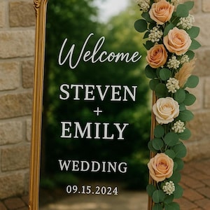 May include: A tall, rectangular mirror with a gold frame featuring white text that reads Welcome STEVEN + EMILY WEDDING 09.15.2024. The right side of the mirror is decorated with a vertical arrangement of peach-colored roses, green eucalyptus leaves, and small white filler flowers.