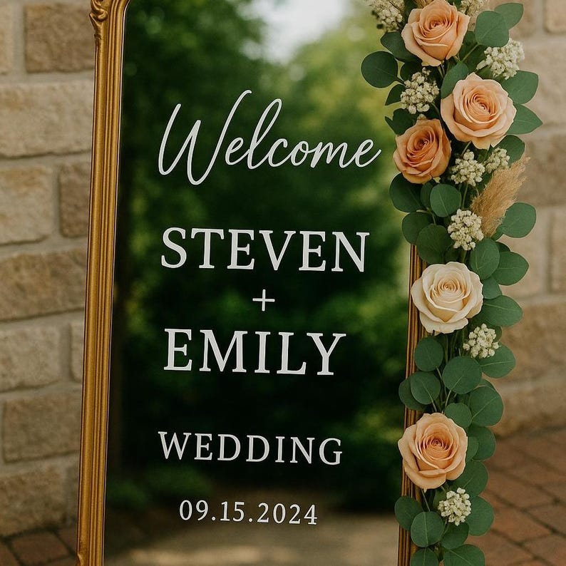 May include: A tall, rectangular mirror with a gold frame featuring white text that reads Welcome STEVEN + EMILY WEDDING 09.15.2024. The right side of the mirror is decorated with a vertical arrangement of peach-colored roses, green eucalyptus leaves, and small white filler flowers.