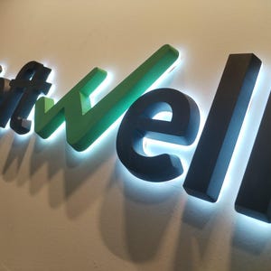 May include: The word "Softwell" is displayed on a wall with illuminated, three-dimensional letters. The letters are in a combination of black and green, with a bright white light outlining each letter, creating a modern and professional aesthetic.