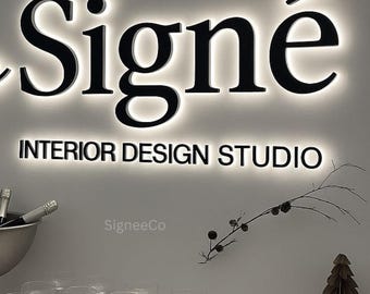 May include: A wall-mounted sign with backlit black lettering reading Signé INTERIOR DESIGN STUDIO. Below the sign are clear glass coupes, a silver bowl with bottle necks, a small brown candle holder, and a paper tree decoration.