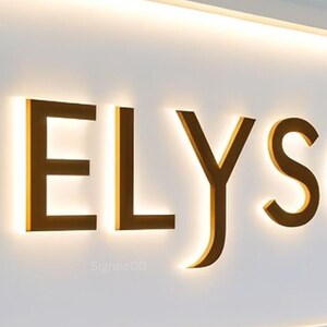 以下が含まれることがあります： 白い壁に設置された、温かみのあるゴールドカラーで「ELYS」と書かれた照明付きのサイン。文字はバックライトで照らされ、光る効果を生み出しています。このサインは、ビジネスや施設用である可能性が高いです。「SigneeCO」の文字が下に表示されています。