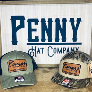 Peut inclure: Deux casquettes de baseball avec le texte "Cougar Hunting Society Mounting Trophies Only" sur un patch en cuir. L'une est vert olive avec un dos en filet noir, l'autre est camouflage avec un patch beige.