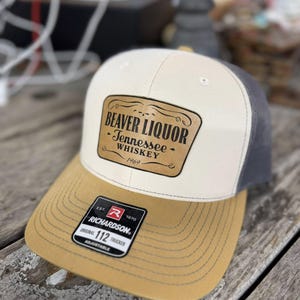 Peut inclure: Casquette de camionneur beige, grise et beige avec le texte "BEAVER LIQUOR - Tennessee Whiskey 1969" sur un écusson marron. La casquette a une visière incurvée et une sangle réglable.
