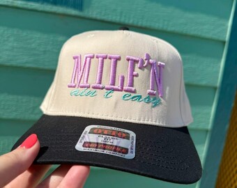 May include: A beige and black baseball cap with the text "MILF'N ain't easy" embroidered in purple and teal. The cap has a black brim and a classic fit.