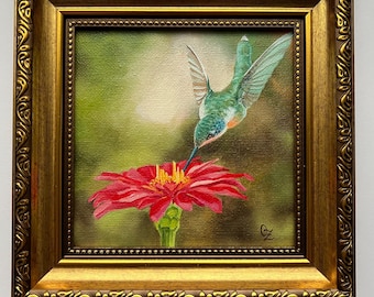 May include: A framed painting of a hummingbird hovering near a red flower. The bird has green and blue feathers, and the flower has a yellow center. The frame is gold-colored with ornate detailing.
