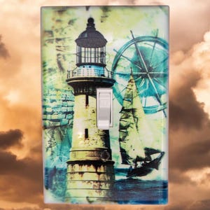 May include: A light switch cover with a nautical theme featuring a lighthouse, a sailboat, and a compass wheel. The background is a watercolor-style design in shades of blue, green, and yellow.