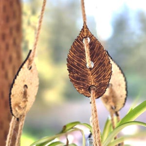 May include: Close-up of hanging leaf-shaped wooden ornaments. The ornaments are brown with detailed leaf vein patterns and are suspended by natural fiber rope. The background is blurred, with a glimpse of greenery.