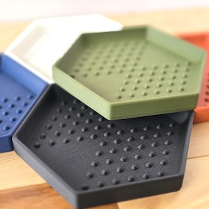 May include: A collection of hexagonal trays in various colors, including blue, white, green, black, and orange. Each tray has a textured surface with small, raised dots. The trays are arranged in a close-up shot, showcasing their design.