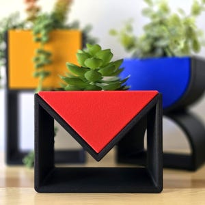 May include: Geometric planters in primary colours. A red triangle planter sits in front of a yellow square and a blue semi-circle planter. The planters are black with colourful accents and hold small green plants. Modern design.