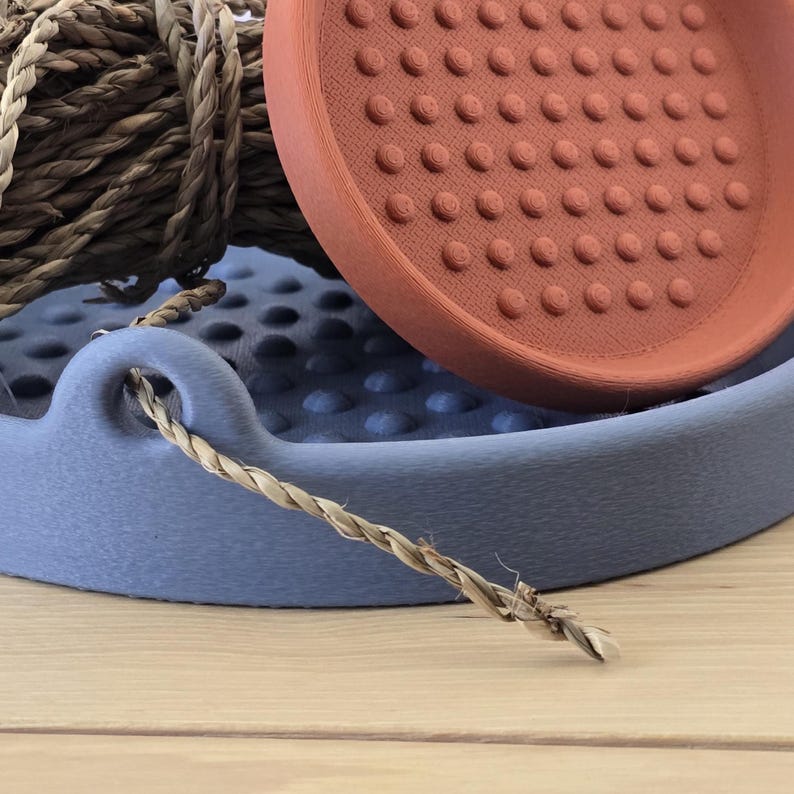 May include: A close-up shot of a blue tray with a woven rope handle and a terracotta-coloured, textured plate. A bundle of natural rope sits behind the plate. The objects are on a light wood surface.