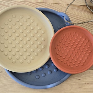 May include: Three ceramic plant saucers in different sizes and colours. The largest is light beige, the medium is terracotta orange, and the base is a muted blue. Each saucer has a textured, dotted surface.
