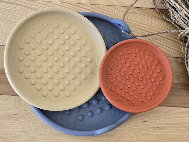 May include: Three ceramic plant saucers in different sizes and colours. The largest is light beige, the medium is terracotta orange, and the base is a muted blue. Each saucer has a textured, dotted surface.