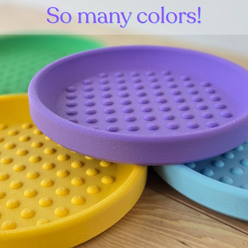May include: Four brightly coloured, round trays are shown in a close-up. The trays are yellow, purple, green, and blue. Each tray has a textured surface with evenly spaced, raised dots. The text "So many colours!" is at the top.
