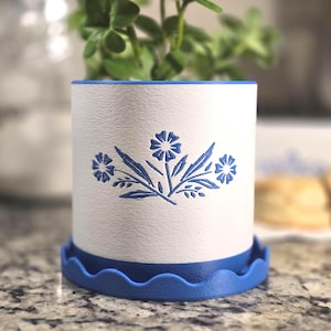 May include: A white ceramic planter with a blue floral design and a matching blue scalloped base. The planter holds a small green plant. The design is reminiscent of vintage kitchenware.
