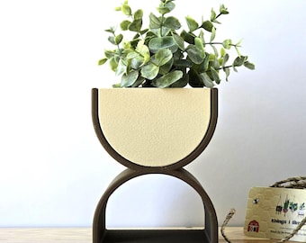 May include: A decorative, two-tiered planter with a dark brown wooden frame and a cream-colored interior. The top section holds a green leafy plant. The planter has a unique, modern design, perfect for displaying small plants or succulents.