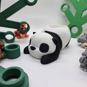 Puede incluir: Un macetero de panda blanco y negro con cara de dormir. El macetero está hecho de plástico impreso en 3D y tiene una pequeña abertura en la parte superior para plantar.