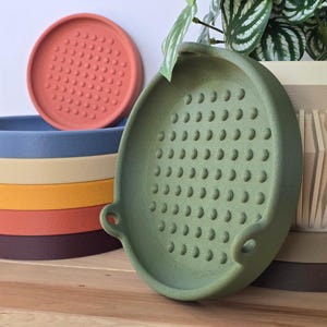 May include: A collection of colourful plant saucers in shades of red, blue, beige, yellow, orange, and burgundy. A green saucer with handles and a perforated surface is in the foreground. A red saucer with a dotted pattern is in the background.
