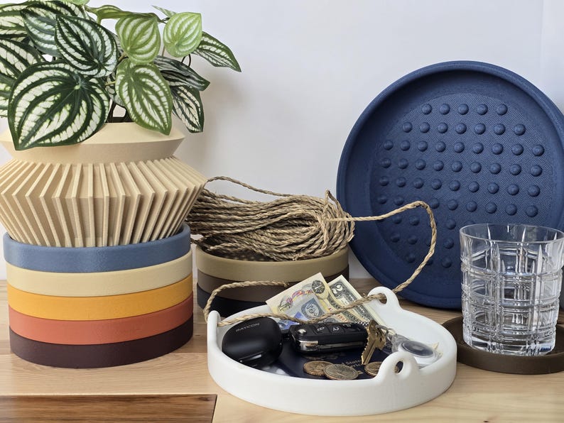 May include: A selection of colourful trays and containers. A white tray holds keys, money, and a car key. A blue tray with a dotted pattern sits next to a clear glass. A plant sits in a beige, geometric vase.
