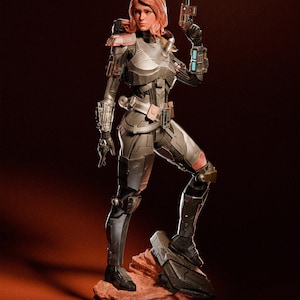 Könnte beinhalten: Eine weibliche Actionfigur, die einen grauen und schwarzen futuristischen Anzug mit einem roten und schwarzen Blaster in der rechten Hand trägt. Die Figur steht auf einem felsigen Sockel.