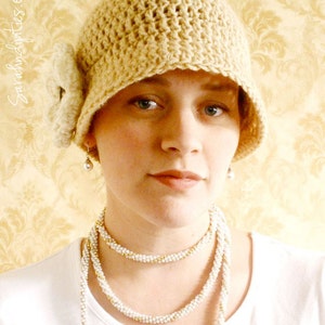 May include: A beige crocheted cloche hat with a floral accent. The hat is paired with a layered pearl necklace with gold accents. The background is a patterned, light yellow wallpaper.